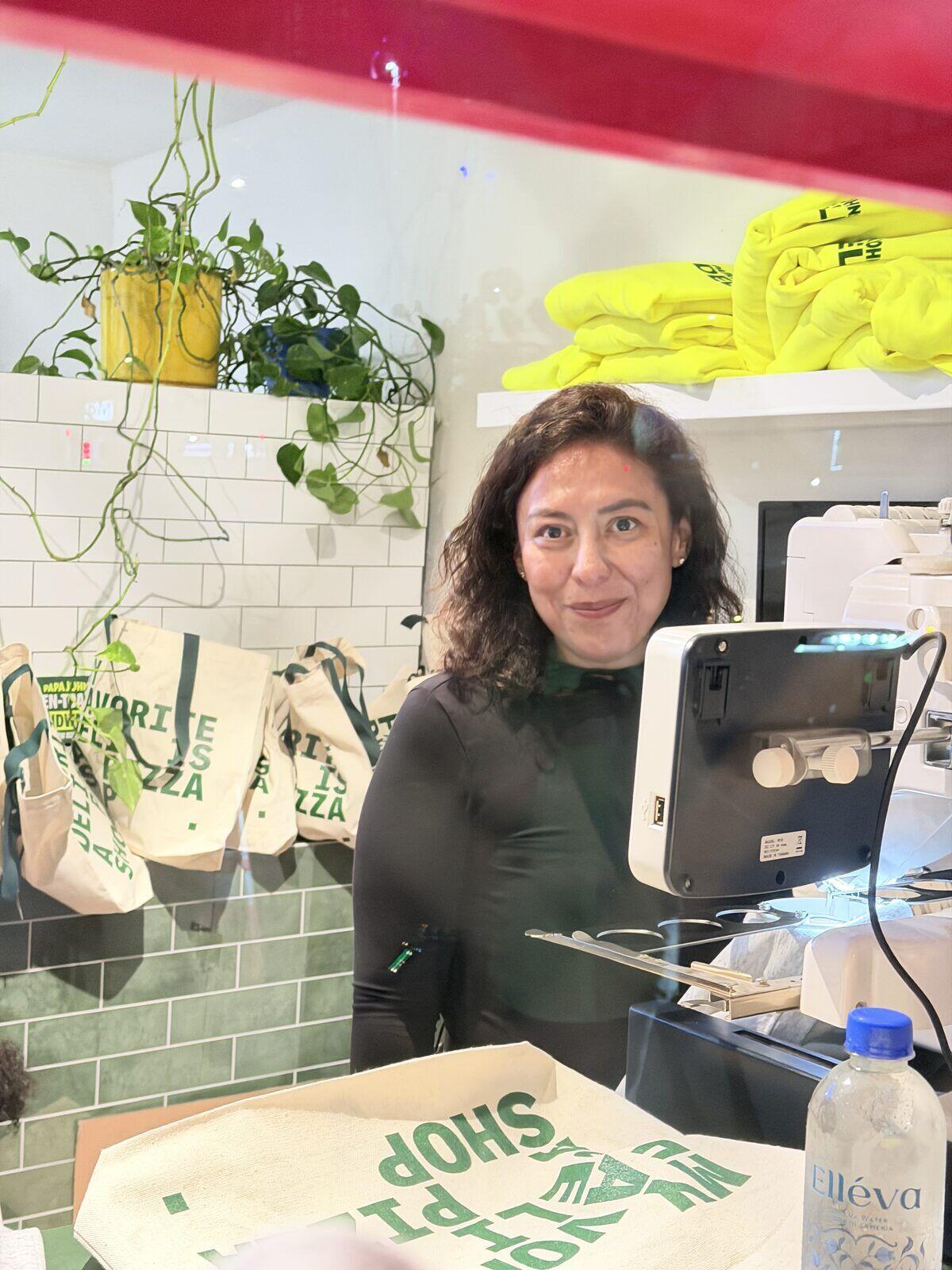 A Merch Troop crew member at a live embroidery machine with stitched totes and neon hoodies on the rack behind
